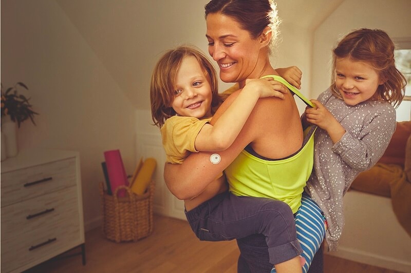 Frau trägt Kinder auf dem Arm und FreeStyle Libre 3 Sensor
