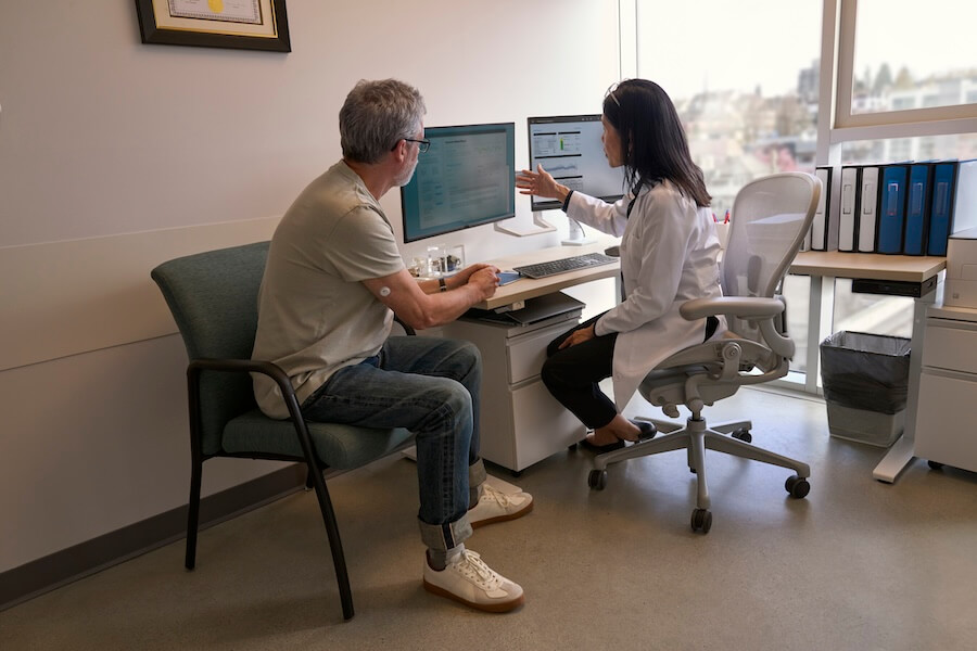 Mann mit FreeStyle Libre Sensor am Arm sitzt neben einer Ärztin. Beide schauen auf einen Computer mit LibreView Berichten